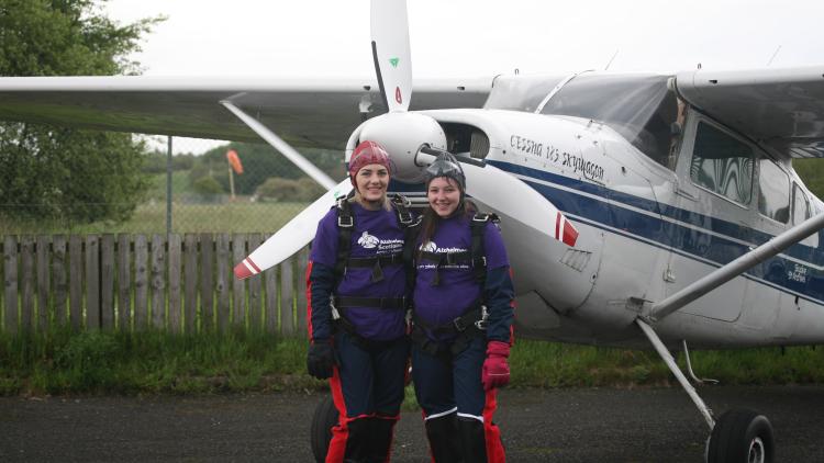Two skydivers with Purple Alzheimer Scotland t-shirts in front of a small plane and in skydiving safety equiptment