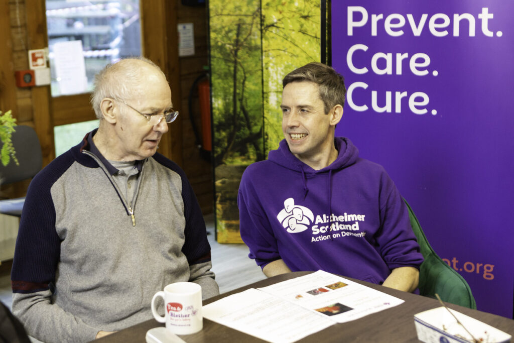 A man talks to an Alzheimer Scotland member of staff wearing a purple branded hoodie