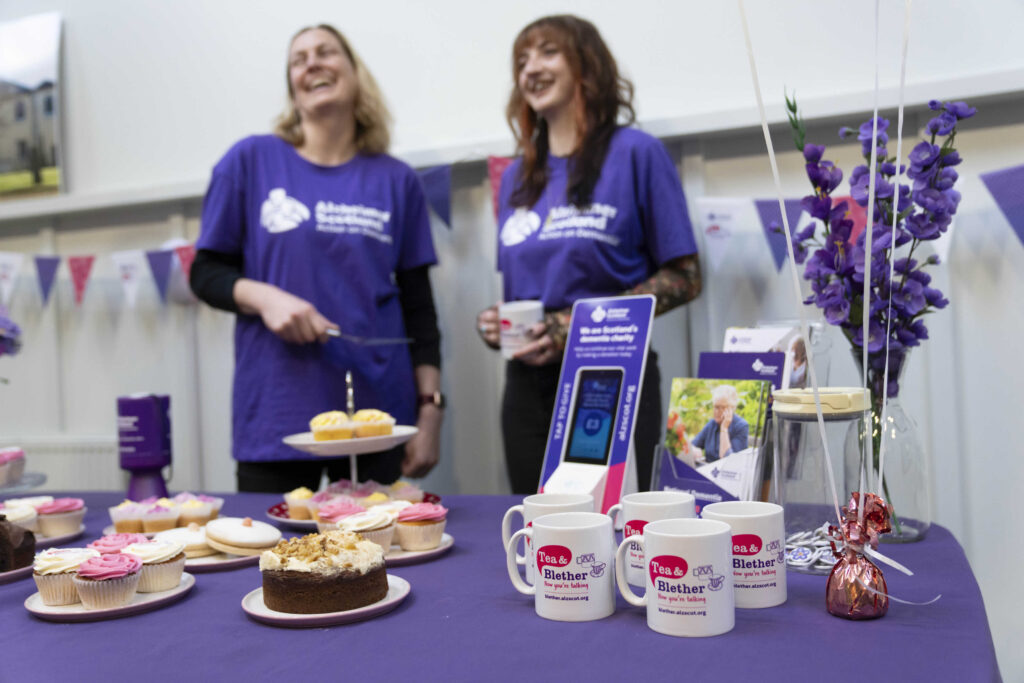 Tea and belther mugs and cakes on a purple table with Alzheimer Scotland staff in background