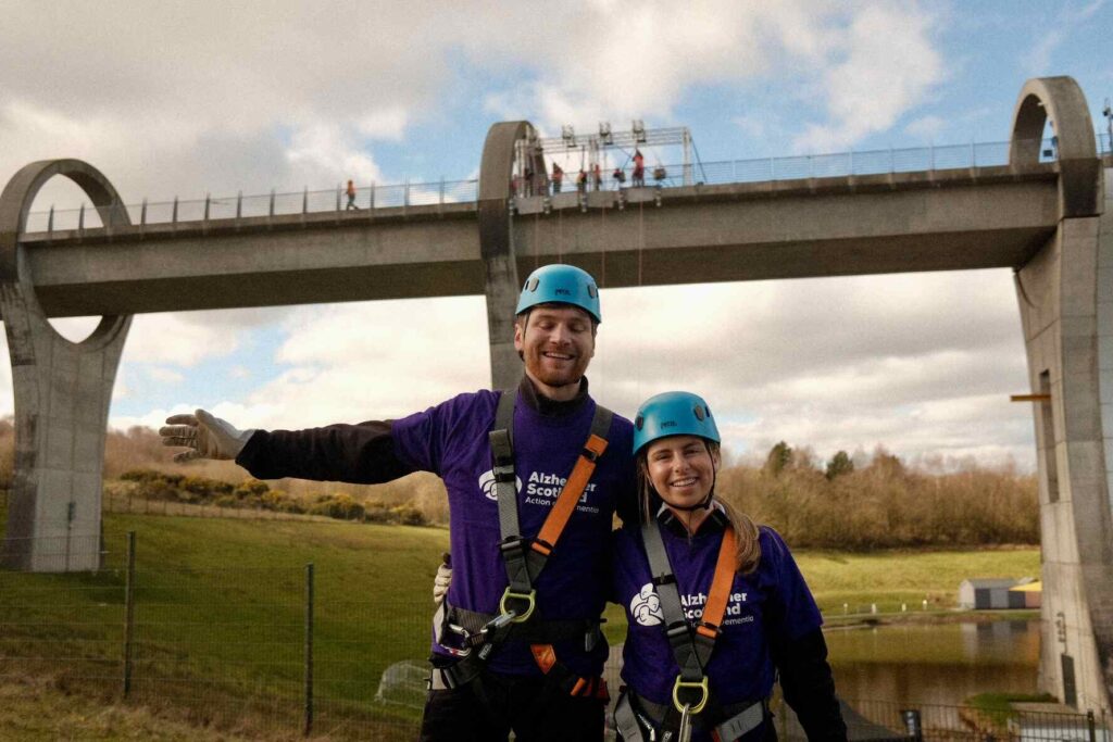 Two people standing together with abseil safety equiptment on with Falkirk wheel behind them