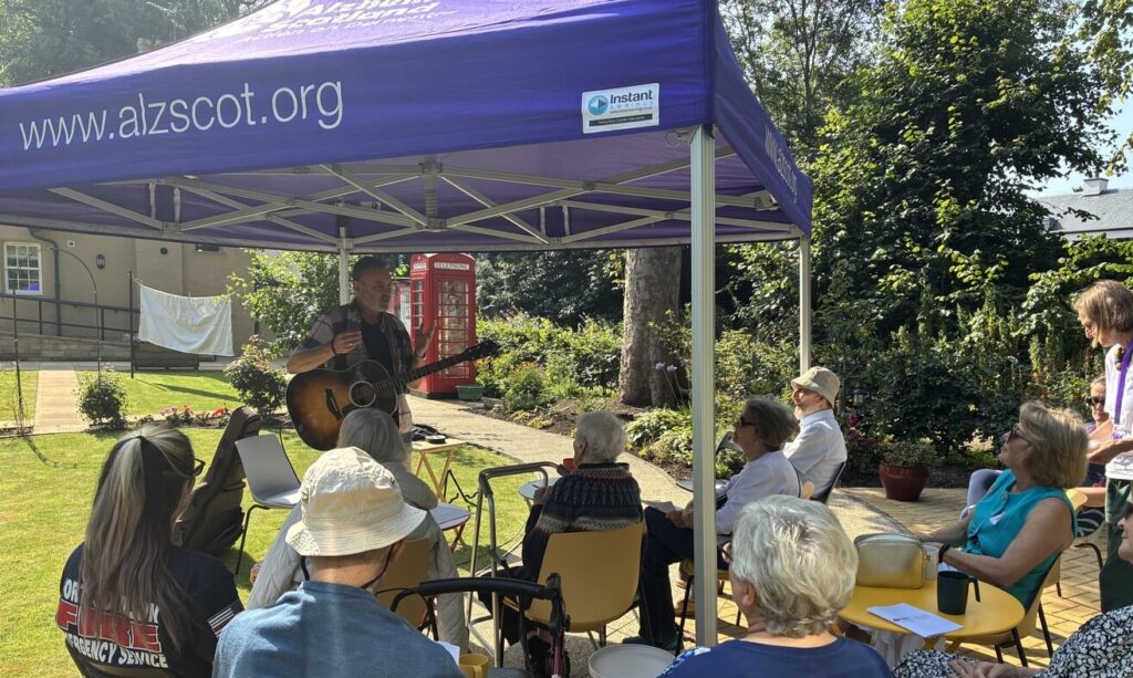 A man plays guitar to a crowd under a purple gazeebo